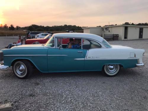 A classic two-tone vintage car parked on gravel, with a sunset in the background.