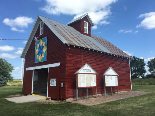 Red barn with a colorful quilt pattern on the side, surrounded by green grass and a blue sky with clouds.