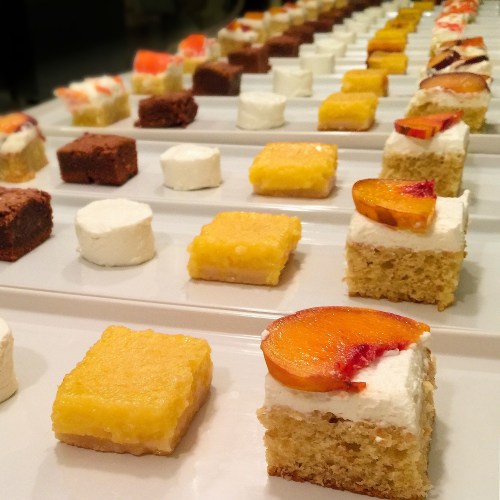 A variety of small desserts arranged in rows, including cakes, marshmallows, and fruit-topped treats on white plates.