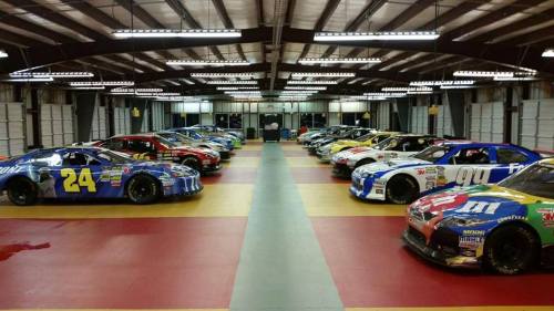 A row of colorful NASCAR race cars lined up in a garage with bright overhead lights and a polished floor.