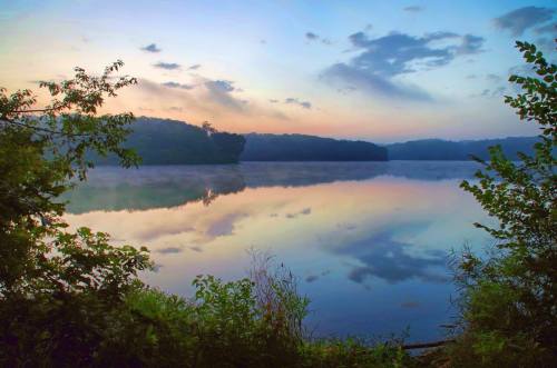 A serene lake at dawn, reflecting colorful clouds and trees, surrounded by mist and greenery.