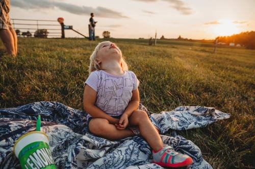 A young child sits on a blanket in a grassy field, looking up at the sunset with a drink beside her.
