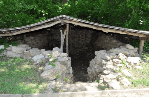 A stone structure with a wooden roof, partially surrounded by greenery, resembling a small shelter or ruins.