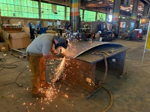 A worker in a workshop welds metal, creating sparks, with machinery and tools visible in the background.