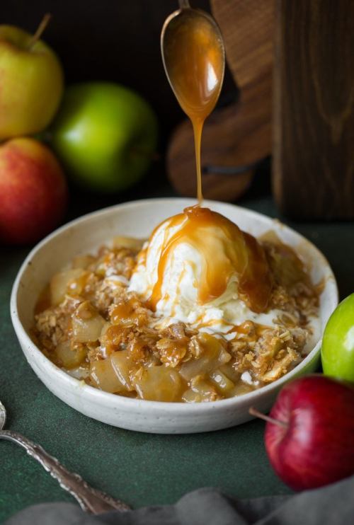 A bowl of warm apple crisp topped with vanilla ice cream and drizzled with caramel, surrounded by fresh apples.