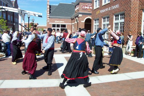 A lively group of dancers in traditional costumes perform in a bustling outdoor plaza, surrounded by an audience.