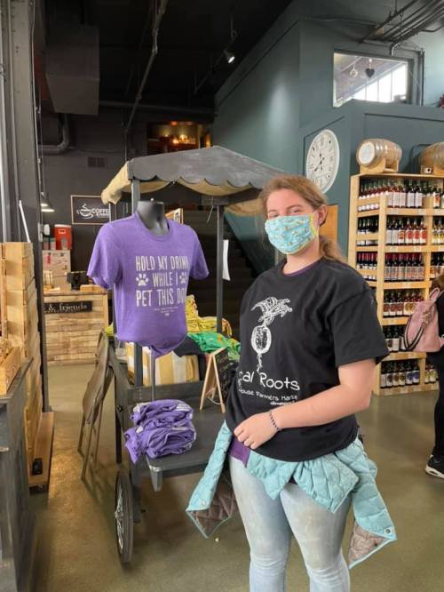 A young woman in a black t-shirt stands near a display of t-shirts in a store, wearing a mask and smiling.