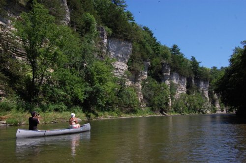 A person in a canoe takes a photo of another person on a river, surrounded by lush greenery and rocky cliffs.