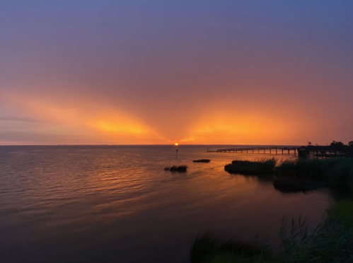 A serene sunset over a calm body of water, with vibrant orange and purple hues and a distant pier.