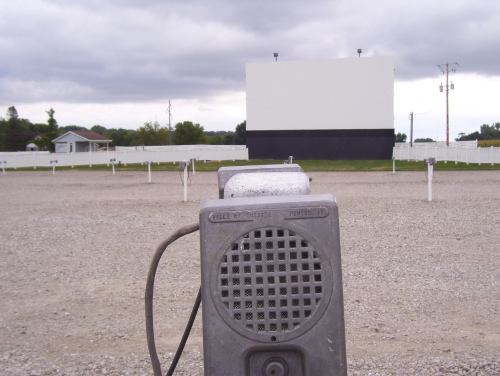 A vintage drive-in movie speaker in the foreground, facing a blank movie screen under a cloudy sky.
