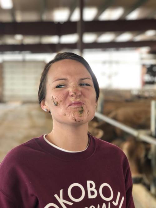 A woman with a playful expression has dirt and grass on her face, standing in a barn with cows in the background.