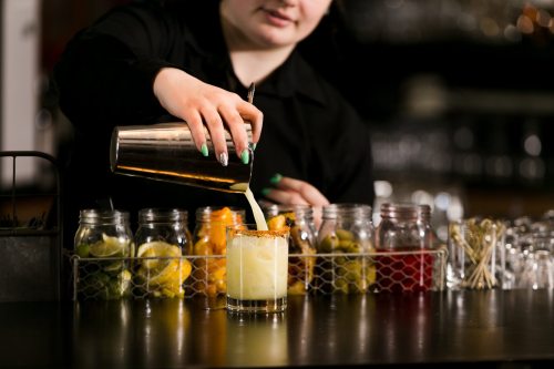 A bartender pours a creamy cocktail into a glass, surrounded by jars of garnishes on the bar.