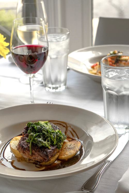 A plate of steak with sautéed greens on potatoes, accompanied by glasses of red wine and water on a dining table.