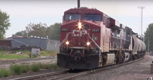 A red and black freight train with the number 8857 approaches on a track, surrounded by greenery and industrial buildings.