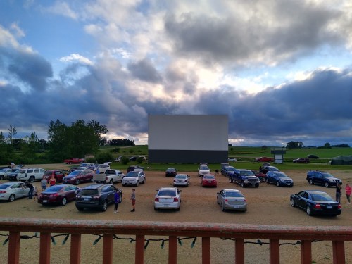 A drive-in movie theater with cars parked in front of a large blank screen under a cloudy sky.