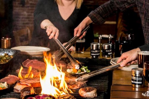 Two people using tongs to grill meat and vegetables over an open flame at a lively dining table.