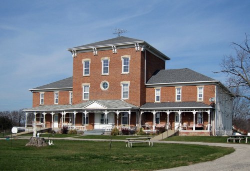 A large, historic brick building with a porch, surrounded by green grass and trees under a blue sky.