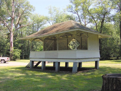 A white gazebo with a sloped roof, situated on a grassy area surrounded by trees.