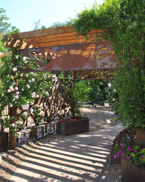 A garden entrance with a wooden arch, adorned with flowers and decorative metalwork, casting shadows on the path.
