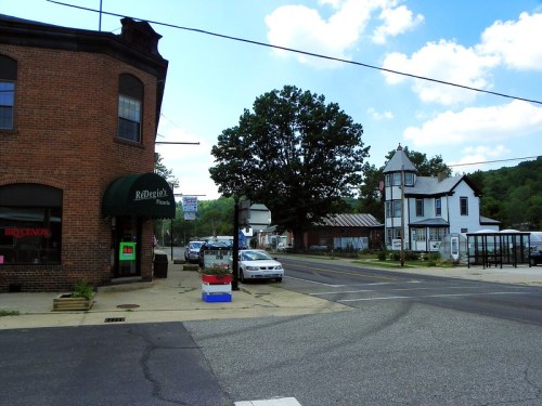 A small town street scene featuring brick buildings, a tree, and parked cars under a blue sky with clouds.