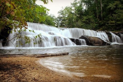 A serene waterfall cascades over rocks into a calm pool, surrounded by lush greenery and sandy shore.