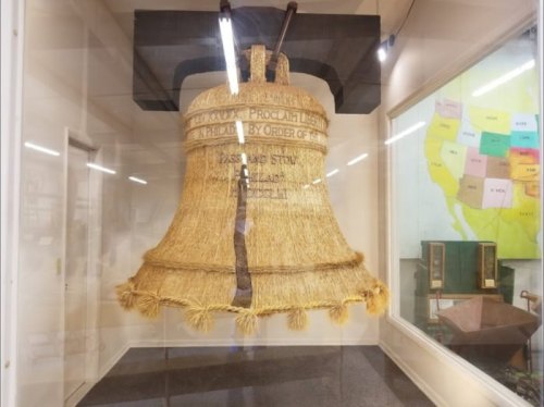 A large decorative bell made of straw displayed in a glass case, with inscriptions on its surface.