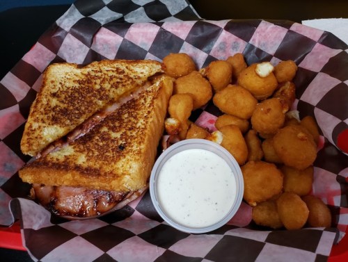 A grilled sandwich with ham and cheese, served with a side of golden corn fritters and a small cup of dipping sauce.