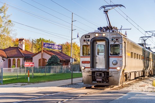 A train approaches a station with a vintage sign, surrounded by trees and a building in the background.