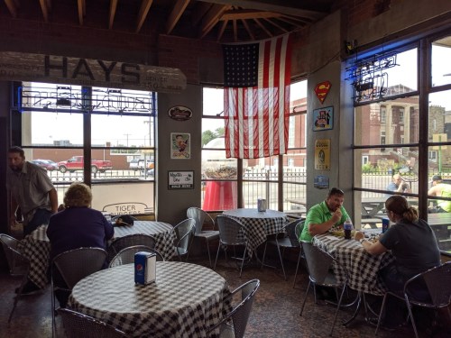 Interior of a diner with checkered tables, an American flag, and patrons seated near large windows.