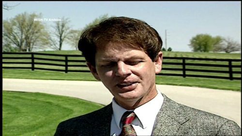 A man in a suit stands outdoors near a fence, with green fields and trees in the background.