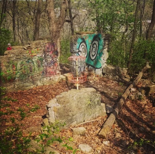 A stone structure in a wooded area, featuring colorful graffiti and a disc golf basket in the center, surrounded by fallen leaves.