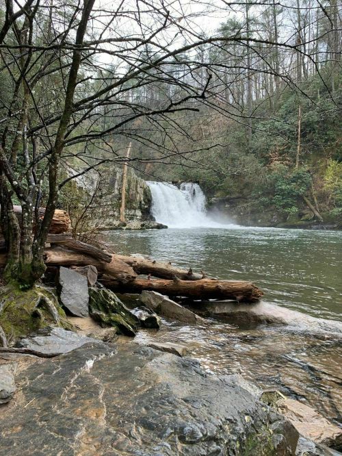 A serene waterfall cascades into a calm river, surrounded by trees and rocky terrain. Mist rises from the water.
