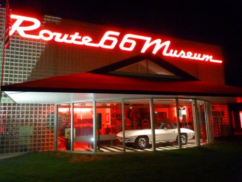 A brightly lit Route 66 Museum at night, featuring a classic car displayed in a glass-fronted area.