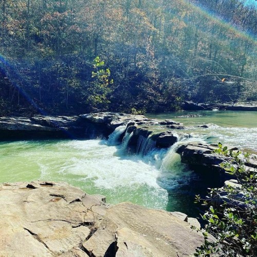 A serene waterfall cascades over rocks into a green pool, surrounded by trees and autumn foliage.