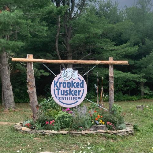 Sign for Krooked Tusker Distillery surrounded by flowers and trees, featuring a cartoon elephant logo.