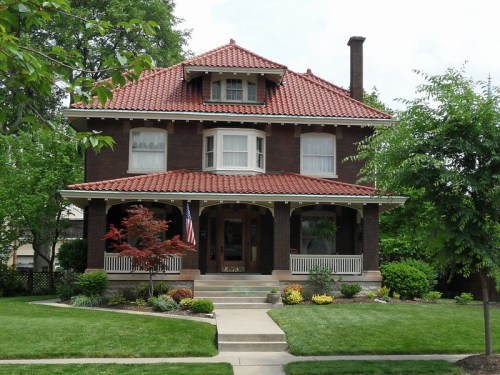 A large, two-story brick house with a red tile roof, surrounded by greenery and a well-maintained lawn.
