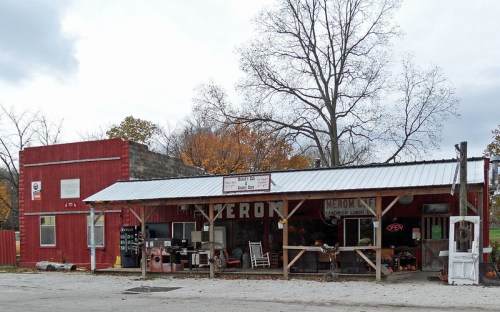 A rustic storefront with a red building, porch seating, and a sign reading "Meron Cafe" under a cloudy sky.