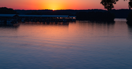 Sunset over a calm lake, reflecting vibrant orange and purple hues, with silhouettes of boat docks in the foreground.