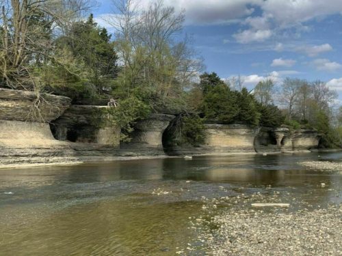 A serene riverbank with layered rock formations and lush greenery under a partly cloudy sky.