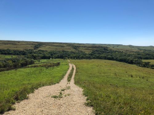 A winding gravel path leads through lush green fields under a clear blue sky. Rolling hills are visible in the distance.