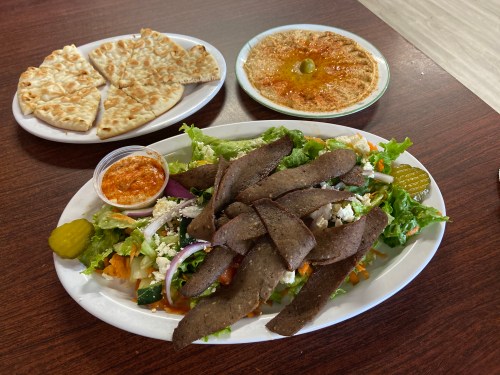 A platter of salad topped with sliced meat, pickles, and a side of hummus and flatbread on a wooden table.