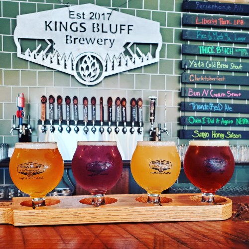 A flight of four craft beers in glasses on a wooden paddle, with a brewery logo and taps in the background.