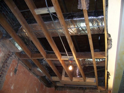 Exposed ceiling with wooden beams, ductwork, and insulation, illuminated by a hanging light bulb.
