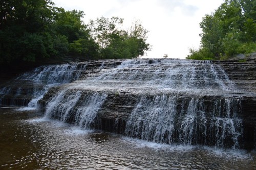 A serene waterfall cascading over rocky ledges, surrounded by lush greenery and a calm pool below.
