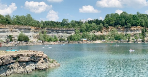 A serene lake surrounded by rocky cliffs and lush greenery under a blue sky with fluffy clouds.