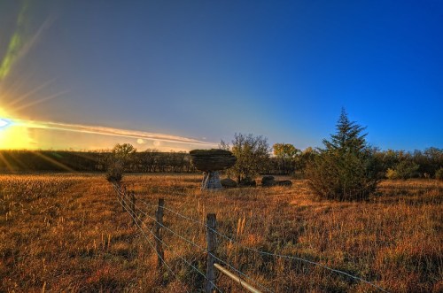 A serene landscape at sunset, featuring a large rock formation, grassy fields, and a clear blue sky.