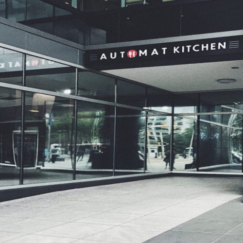 Exterior view of Automat Kitchen with large glass windows reflecting the surrounding cityscape.