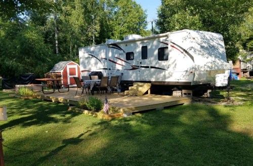 A parked RV on a wooden deck surrounded by grass and trees, with outdoor furniture and a small shed nearby.