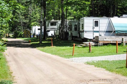 A dirt road lined with RVs and a small shed, surrounded by green trees and grass in a camping area.
