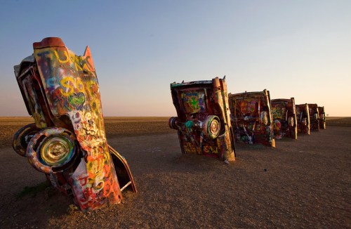 A row of colorful, graffiti-covered cars partially buried in the ground against a clear sky.
